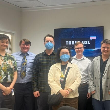 A group of smiling people standing in front of a slide deck title slide reading Trans 101 with a trans flag graphic